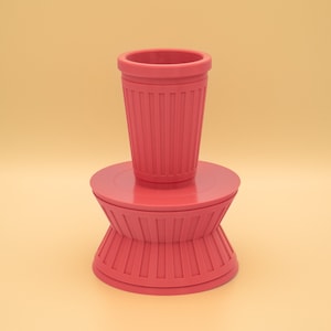 May include: A pink ceramic planter with a fluted design. The planter has a wide base and a narrow top, and it is sitting on a pink pedestal.