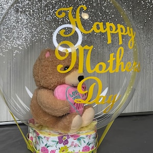 May include: A clear balloon with a yellow ribbon and a teddy bear holding a pink heart inside. The balloon has the words "Happy Mother's Day" written on it in yellow.