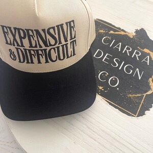 Peut inclure: Casquette de baseball beige et noire avec le texte "EXPENSIVE & DIFFICULT" en noir. La casquette a une visière noire et un bouton noir sur le dessus. Le logo "CIARRA DESIGN CO." est visible en arrière-plan.