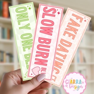 May include: Three rectangular bookmarks with rounded corners. The left is light green with a checkerboard pattern and the text ONLY ONE BED. The center is pink with a heart pattern and SLOW BURN. The right is peach with a checkerboard pattern and FAKE DATING.