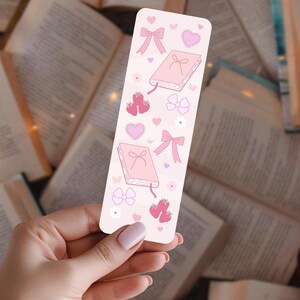 May include: A pink bookmark with illustrations of books, bows, strawberries, hearts, and flowers. The bookmark is held in front of a background of open books.