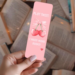 Peut inclure: Un marque-page rose avec le texte "PLOT TWIST: I NEED MORE BOOKS !" et une illustration à l'aquarelle de deux cerises liées par un nœud. Un petit cœur rouge est en bas. Le marque-page est tenu dans une main.