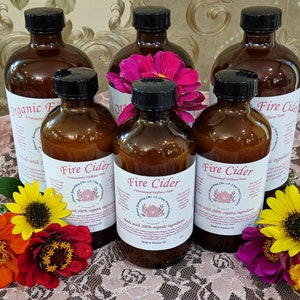 May include: Six amber glass bottles with black lids containing Fire Cider, an unpasteurized vinegar tonic. The bottles are arranged on a white lace tablecloth with pink, yellow, and orange flowers.