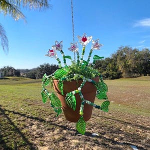 May include: A hanging terracotta pot with a chain, filled with greenery and decorative glass flowers. The flowers have red centers and translucent petals. Green glass leaf and bead strands cascade from the pot, creating a whimsical, nature-inspired design.