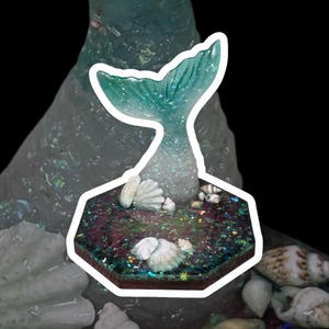 May include: A mermaid tail sculpture with a teal and white color scheme. The tail is set on a hexagonal base with seashells and glitter. The sculpture has a white outline and is set against a dark background.
