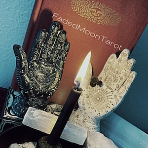 May include: A still life featuring a burning black candle, two decorative hand sculptures, and a book titled "Faded Moon Tarot." The hands are intricately detailed, one black and one white. Crystals and a small bowl are also present.
