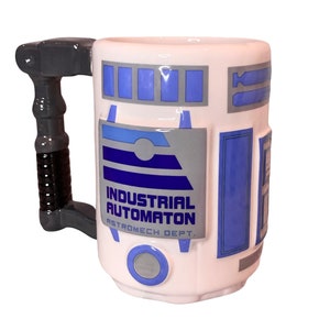 May include: White ceramic mug with a blue and gray R2-D2 design. The mug has a handle shaped like R2-D2's arm. The text on the mug reads "Industrial Automaton Astromech Dept."