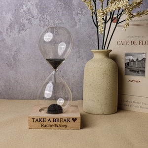 Pode incluir: Um relógio de areia de madeira com areia preta, um símbolo de coração e o texto "TAKE A BREAK Rachel&Joey" na base. O relógio de areia está sobre uma superfície bege com um vaso de flores secas e um livro no fundo.
