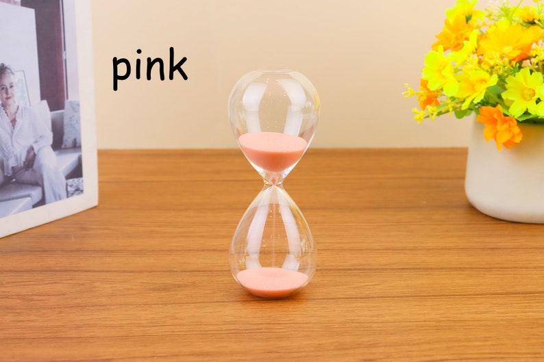 Op de afbeelding: Een roze zandloper met een glazen bol en een heldere zandteller. De zandloper staat op een houten oppervlak.