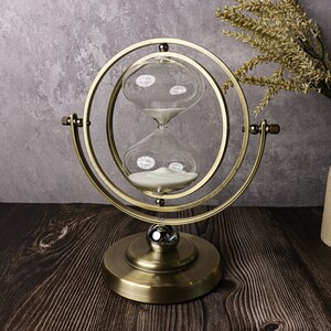 May include: A gold-toned hourglass with white sand inside is mounted on a rotating stand. The stand has a circular base and a ring that holds the hourglass. The hourglass is on a dark wood surface.
