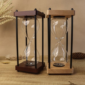 May include: Two hourglasses with clear glass bulbs and wooden frames. One has a dark brown frame, the other a light wood frame. Both are supported by black metal rods. The hourglasses sit on a wooden surface.