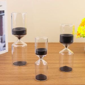 May include: Three glass hourglasses with black sand. The hourglasses are different sizes and are arranged on a wooden surface.