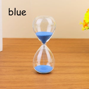 Op de afbeelding: Een blauwe zandloper met zand in de bovenste bol. De zandloper is gemaakt van helder glas en heeft een ronde vorm. Het woord "blauw" is in zwarte letters op een witte achtergrond geschreven.