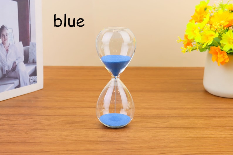 Op de afbeelding: Een blauwe zandloper met zand in de bovenste bol. De zandloper is gemaakt van helder glas en heeft een ronde vorm. Het woord "blauw" is in zwarte letters op een witte achtergrond geschreven.