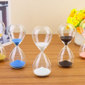 May include: Five glass hourglasses with different colored sand. The hourglasses are on a wooden surface. The sand colors are gold, blue, white, black, and pink.