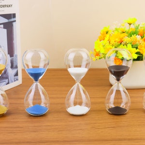 Op de afbeelding: Vijf glazen zandlopers met verschillende gekleurde zand. De zandlopers staan op een houten oppervlak. De zandkleuren zijn geel, blauw, wit, zwart en roze.