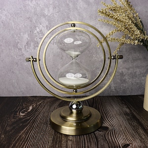 May include: A gold-toned hourglass with a round base and a circular frame. The hourglass is filled with white sand and is set on a dark wood surface.