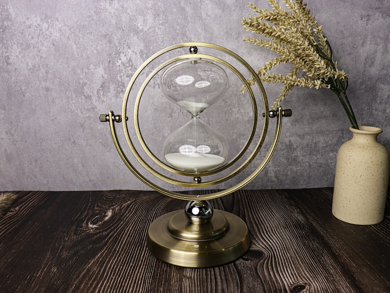 May include: A gold-toned hourglass with a round base and a circular frame. The hourglass is filled with white sand and is set on a dark wood surface.