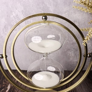 May include: A gold-toned hourglass with a white sand timer inside. The hourglass is suspended in a circular, rotating stand.