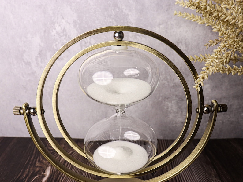 May include: A gold-toned hourglass with a white sand timer inside. The hourglass is suspended in a circular, rotating stand.