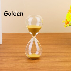Op de afbeelding: Een glazen zandloper met gouden zand. De zandloper staat op een houten oppervlak. Het woord "Golden" is in zwart op een witte achtergrond geschreven.