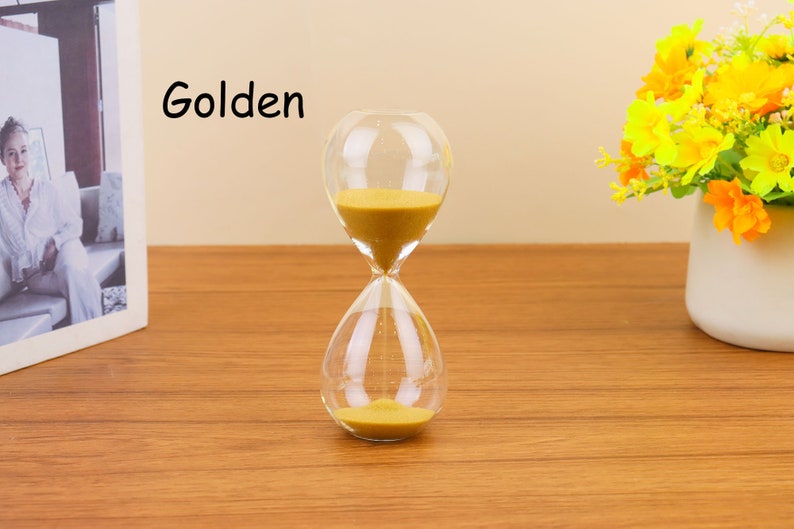Op de afbeelding: Een glazen zandloper met gouden zand. De zandloper staat op een houten oppervlak. Het woord "Golden" is in zwart op een witte achtergrond geschreven.