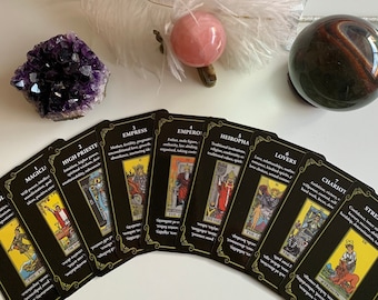 May include: A spread of tarot cards on a white surface with a feather, a pink crystal ball, and a tiger's eye crystal ball. The cards are black with gold trim and feature illustrations of the Major Arcana. The cards are labeled with the numbers 1 through 7 and the names of the cards: Magician, High Priestess, Empress, Emperor, Hierophant, Lovers, and Chariot.
