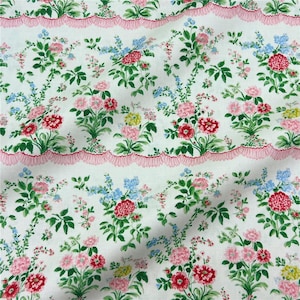 May include: White fabric with a floral pattern featuring pink, red, yellow, and blue flowers. Green leaves and stems are interspersed throughout. A pink scalloped border runs horizontally across the fabric.