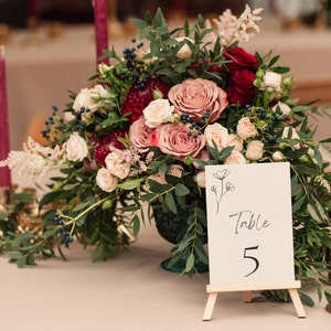 Può includere: Un cartello con il numero del tavolo con il numero 5, con le parole "Table 5" scritte in corsivo. Il cartello è su un piccolo cavalletto ed è circondato da un'arrangiamento floreale di rose rosa e rosse, verde e candele.