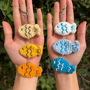 May include: Crocheted fish keychains in yellow, orange, white, and blue. Each fish has a silver keyring attached. The fish are held up against a background of green foliage.