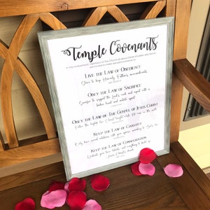 Op de afbeelding: Een ingelijste print met de titel "Temple Covenants" met tekst over religieuze principes. De print staat in een zilveren lijst en rust op een houten bank. Verspreid rond het frame liggen rode en roze rozenblaadjes.