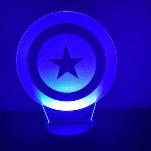 May include: A blue and white Captain America shield shaped night light. The shield has a white star in the center and is illuminated with a blue light.