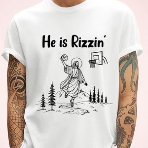 以下が含まれることがあります： 白いTシャツに、バスケットボールをしているイエスの黒いグラフィックと、「He is Rizzin'」というテキストがプリントされています。