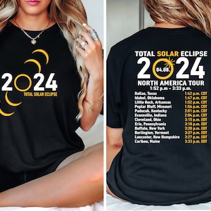 May include: Black t-shirt featuring white and yellow graphics. The front reads "2024 TOTAL SOLAR ECLIPSE" with a crescent and circle design. The back lists a North America Tour with cities and times.