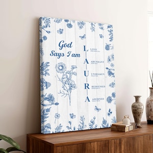 May include: A framed canvas print with a white wood background and blue floral illustrations. The text "God Says I Am" is at the top, with "LAURA" spelled out vertically, each letter associated with a positive affirmation.