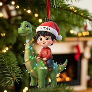 May include: A Christmas ornament featuring a cartoon boy riding a green dinosaur adorned with string lights. The boy wears a red sweater and Santa hat with the name "LUCAS". The year "2025" is displayed on the dinosaur.