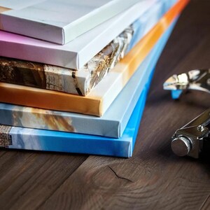 May include: A stack of canvas prints with various images, including a beach scene, a cityscape, and a landscape. The prints are stacked on a wooden surface, and a stapler is visible in the foreground.