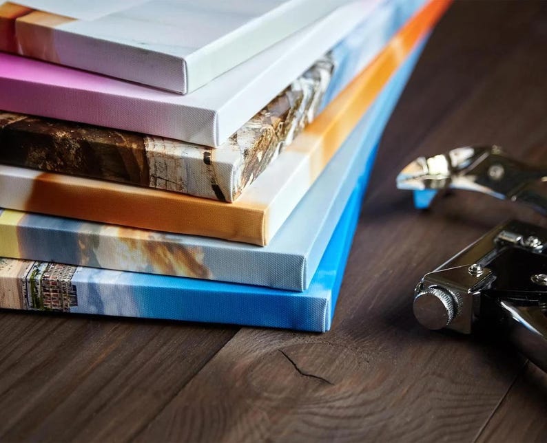 May include: A stack of canvas prints with various images, including a beach scene, a cityscape, and a landscape. The prints are stacked on a wooden surface, and a stapler is visible in the foreground.
