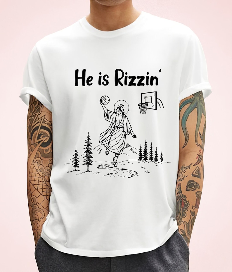 以下が含まれることがあります： 白いTシャツに、バスケットボールをしているイエスの黒いグラフィックと、「He is Rizzin'」というテキストがプリントされています。