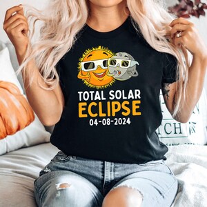 May include: Black t-shirt featuring a cartoon sun and moon wearing sunglasses. The text reads "Total Solar Eclipse 04-08-2024".