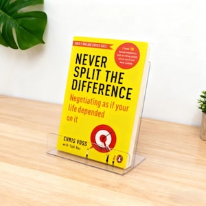 Könnte beinhalten: Ein klarer Acryl-Buchständer mit dem Buch "Never Split the Difference" von Chris Voss. Das Buch hat einen gelben Einband mit schwarzem Text und einer roten Zielscheibe. Der Ständer steht auf einer hellen Holzoberfläche.
