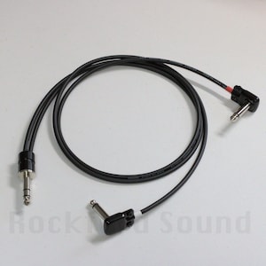 May include: Black instrument cable with two 1/4 inch (6.35 mm) connectors. One connector is straight, and the other is angled. The cable is coiled and lies on a white surface. The cable is for musical instruments.