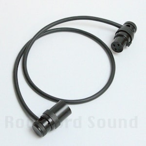 May include: A black XLR cable with a male and female connector. The cable is coiled and lies on a white surface. The connectors have a knurled texture for grip.