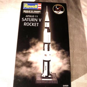 May include: A Revell Apollo 11 Saturn V rocket model kit box featuring a black background with a white rocket illustration, the text Apollo 11 Saturn V Rocket, and the number 04909. The box displays a circular Apollo 11 mission emblem.