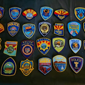 May include: A collection of twenty-four embroidered patches from various Arizona law enforcement agencies, including police, marshal, and sheriff departments. Each patch features unique regional imagery, text, and symbols arranged in a grid pattern.
