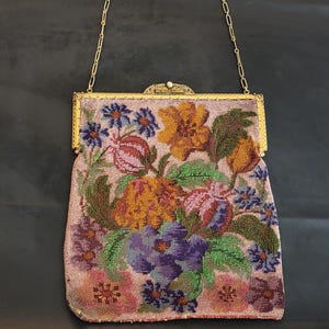 May include: A vintage beaded handbag with a gold-tone frame and chain handle. The bag features a floral design in shades of pink, orange, purple, and green. The clasp has a small pearl accent. The bag is likely from the early 20th century.