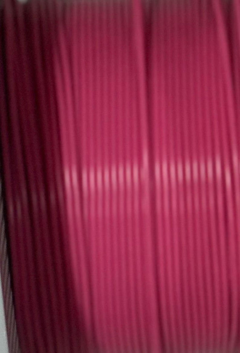 Puede incluir: Una bobina de cord&oacute;n magenta con una textura estriada, cil&iacute;ndrica y consistente. El cord&oacute;n est&aacute; enrollado firmemente en orientaci&oacute;n vertical, mostrando un color rosa intenso uniforme a lo largo de toda la longitud del material visible.