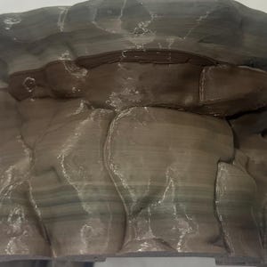 Puede incluir: Un objeto impreso en 3D que presenta una formaci&oacute;n rocosa estratificada con un acabado mate y suave. La estructura muestra estr&iacute;as horizontales en tonos marr&oacute;n oscuro, gris y beige apagado, imitando estratos geol&oacute;gicos naturales con grietas.
