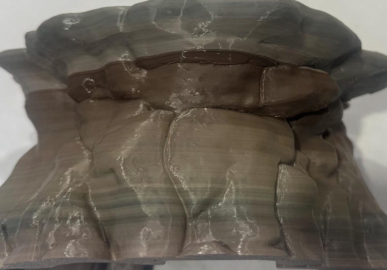 Puede incluir: Un objeto impreso en 3D que presenta una formaci&oacute;n rocosa estratificada con un acabado mate y suave. La estructura muestra estr&iacute;as horizontales en tonos marr&oacute;n oscuro, gris y beige apagado, imitando estratos geol&oacute;gicos naturales con grietas.
