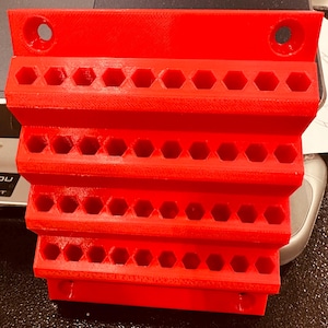 May include: A bright red, 3D-printed tool organizer with multiple rows of hexagonal holes. The organizer has a stepped design and two mounting holes at the top. It is designed to hold various tools.